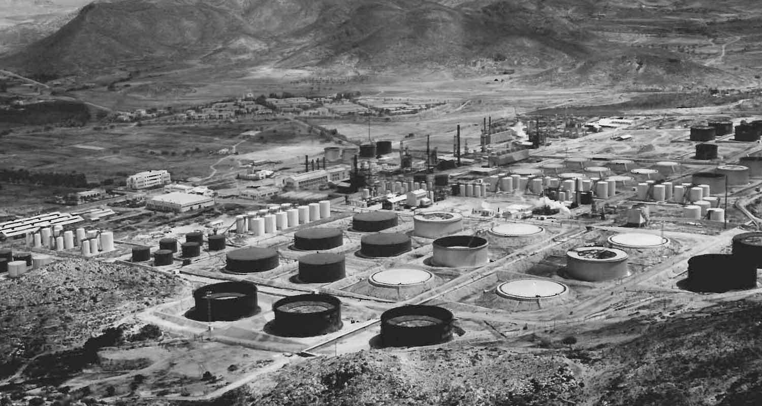 Foto antigua en blanco y negro donde se ve una refinería con tanques negros 