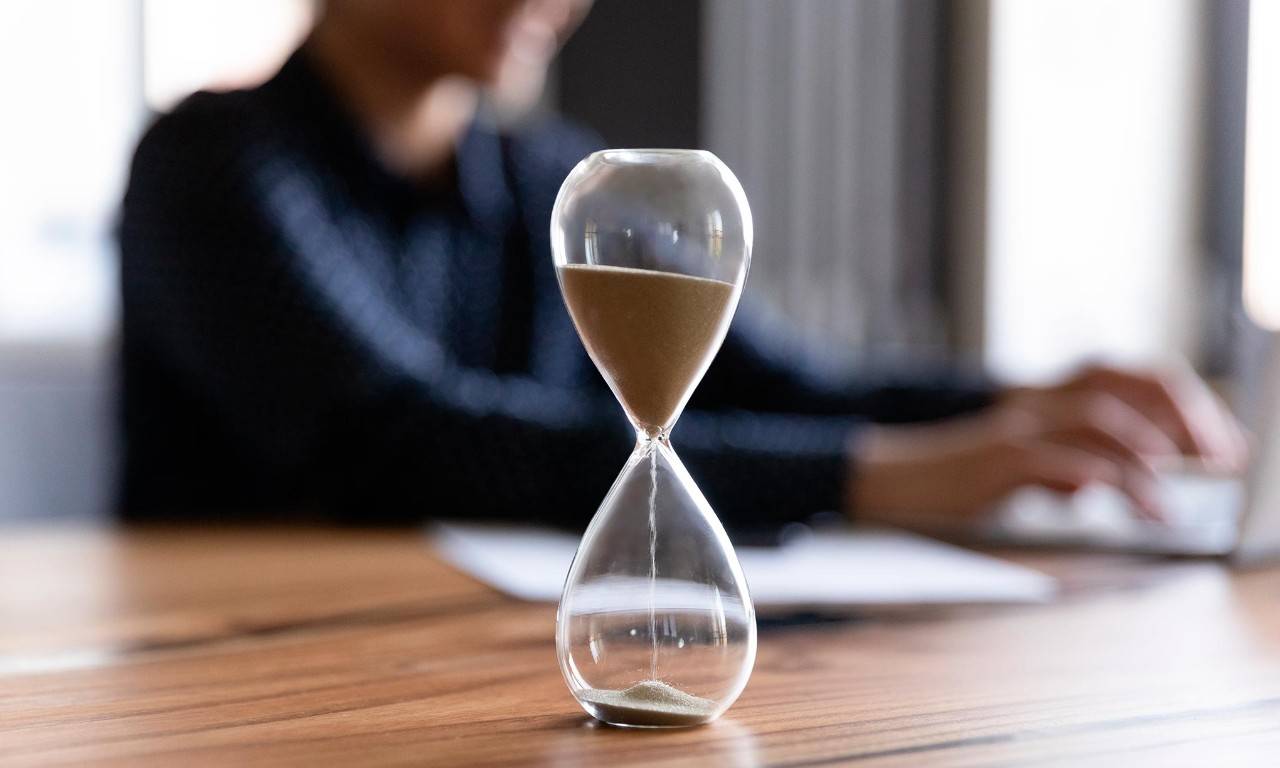 Un reloj de arena en una mesa sobre la que trabaja una persona que gestiona su tiempo