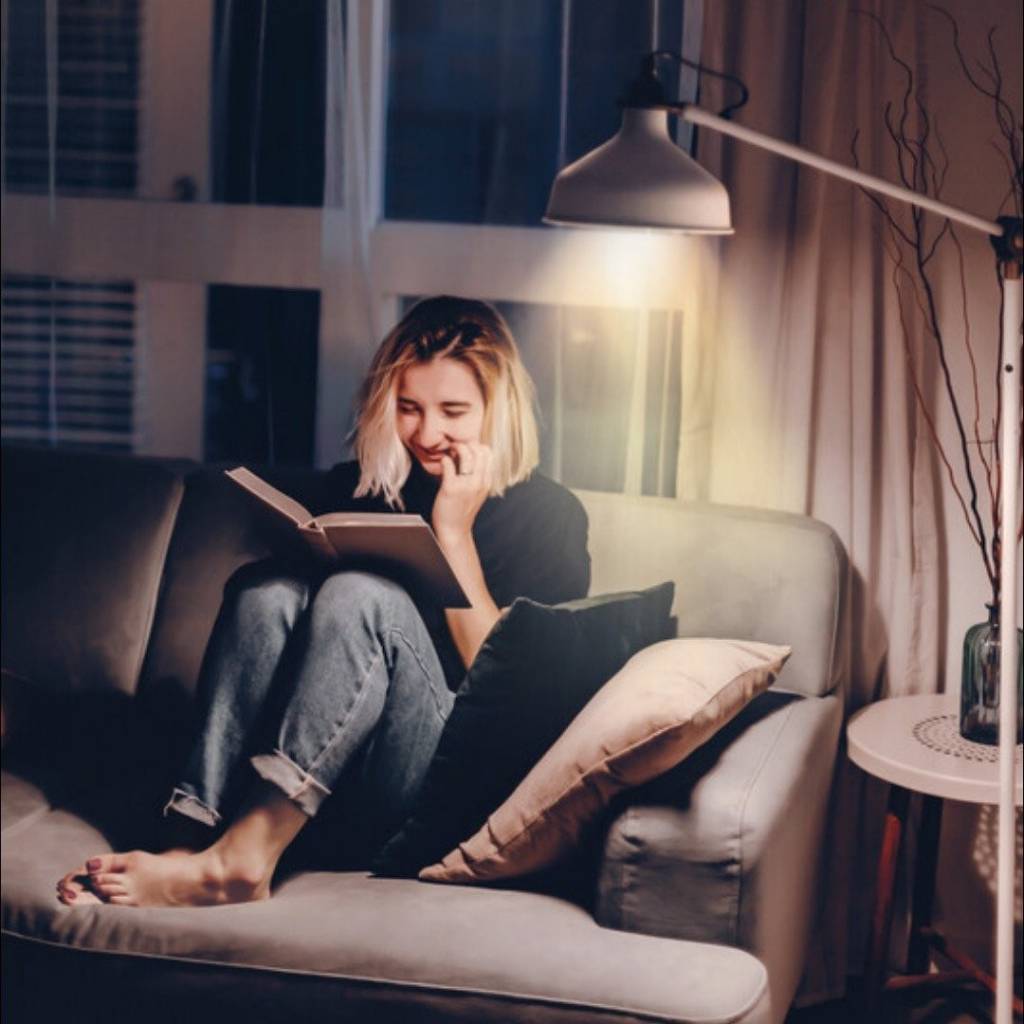 Mujer joven en el sofá leyendo con lámpara al lado